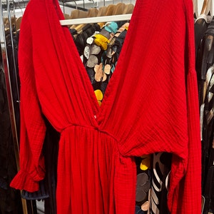 May include: A vibrant red dress with a deep V-neck and long sleeves, hanging on a white hanger. The dress features a cinched waist and a flowing skirt. The fabric appears to be textured. Other clothing items are visible in the background.