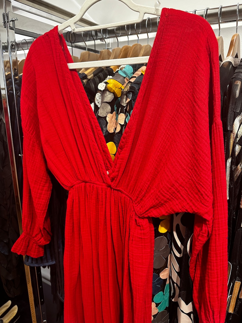 May include: A vibrant red dress with a deep V-neck and long sleeves, hanging on a white hanger. The dress features a cinched waist and a flowing skirt. The fabric appears to be textured. Other clothing items are visible in the background.