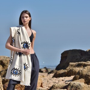 May include: A white beach towel with blue and black embroidered eyes. The towel is draped over a person standing on a sandy beach.