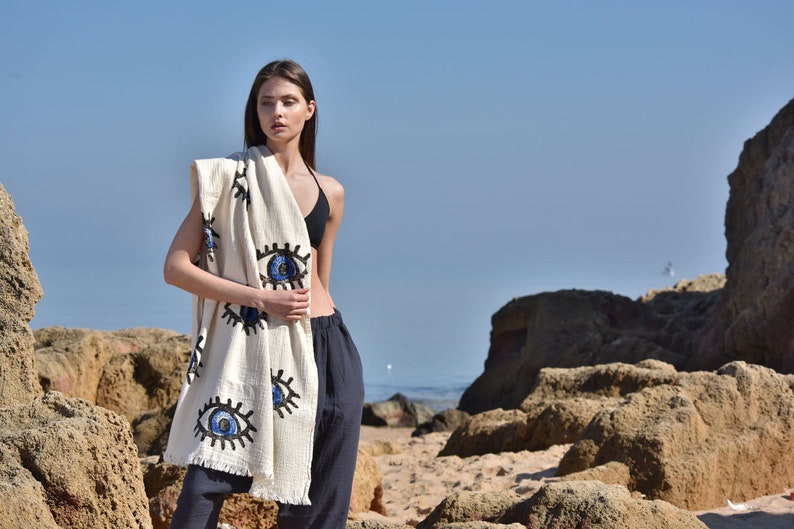 May include: A white beach towel with blue and black embroidered eyes. The towel is draped over a person standing on a sandy beach.