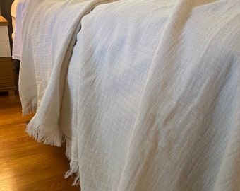 May include: A white, fringed throw blanket draped over a bed with white and gray pillows.