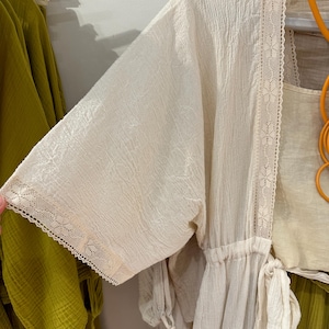 Op de afbeelding: Een witte katoenen badjas met kantversiering en een trekkoord in de taille. De badjas is licht en doorschijnend, waardoor hij perfect is voor warm weer.