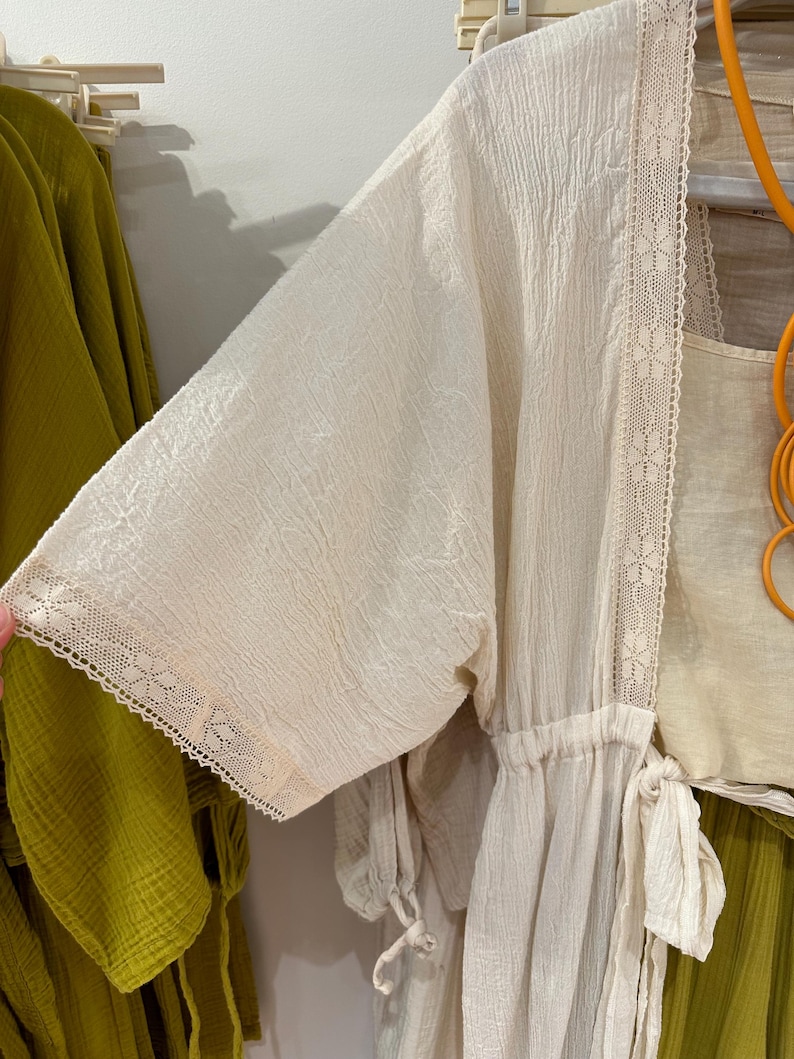 Op de afbeelding: Een witte katoenen badjas met kantversiering en een trekkoord in de taille. De badjas is licht en doorschijnend, waardoor hij perfect is voor warm weer.