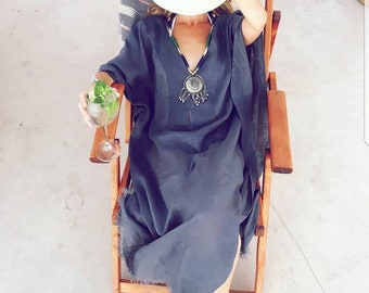 May include: A woman wearing a gray linen kaftan with a silver pendant necklace and a white wide-brimmed hat sits in a wooden chair. She is holding a glass of a drink with a sprig of mint.