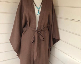 May include: A brown linen kimono with a long, flowing silhouette and wide sleeves. The kimono has a sash that ties at the waist and a deep V-neckline. A turquoise necklace with a coral pendant is worn over the kimono.