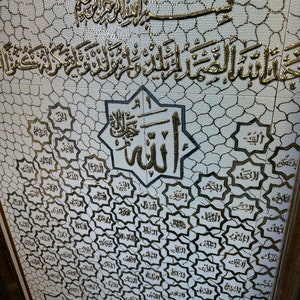 May include: A white ceramic tile with a gold-colored Islamic calligraphy design. The design features the 99 names of Allah in Arabic script, surrounded by a decorative border. The tile is framed in a brown wooden frame.