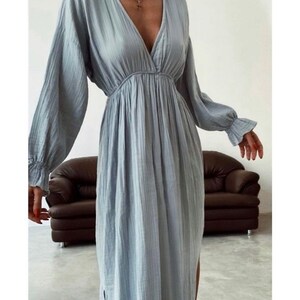 May include: A light blue dress with long sleeves and a V-neck. The dress has a gathered waist and a flowing skirt with a side slit. The sleeves have ruffled cuffs. A gold necklace with a cross pendant is visible.