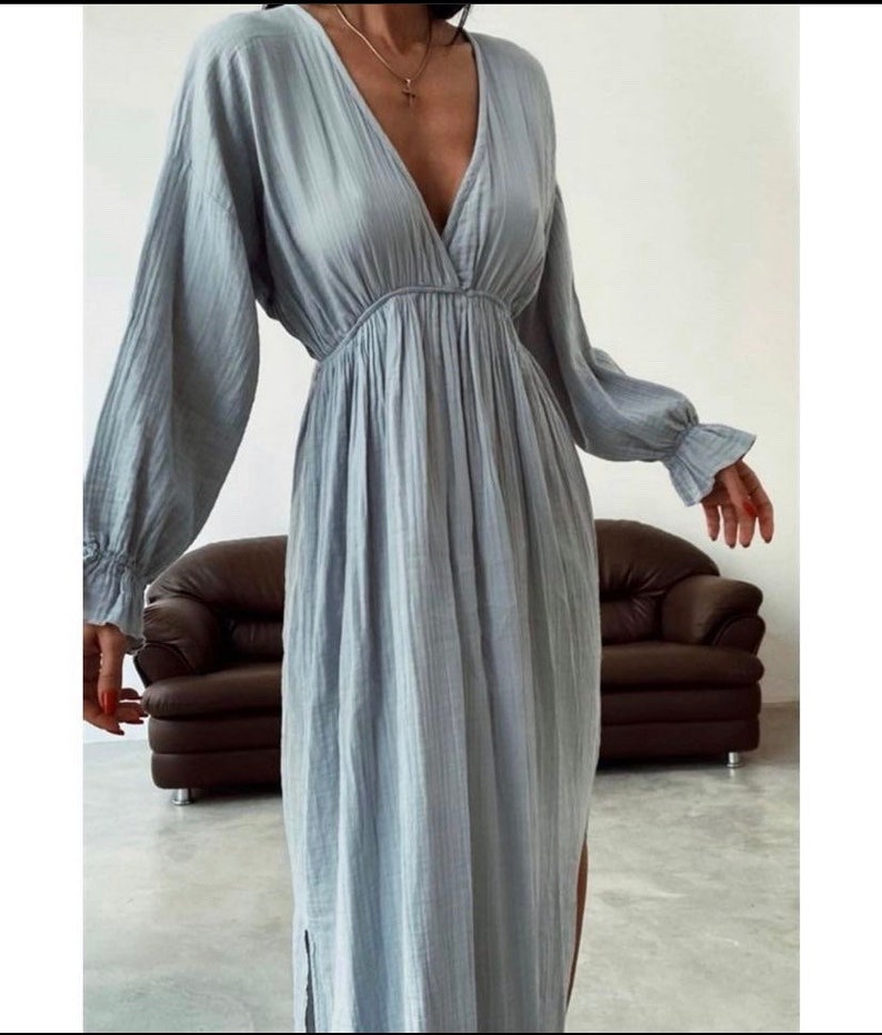 May include: A light blue dress with long sleeves and a V-neck. The dress has a gathered waist and a flowing skirt with a side slit. The sleeves have ruffled cuffs. A gold necklace with a cross pendant is visible.