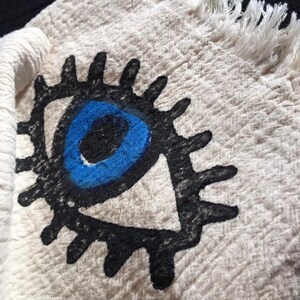 May include: A white cotton towel with a black and blue evil eye design. The towel has a fringed edge.