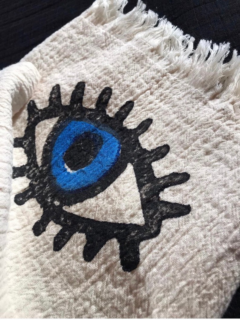 May include: A white cotton towel with a black and blue evil eye design. The towel has a fringed edge.