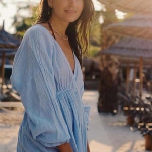 May include: A light blue, long-sleeved dress with a V-neck. The dress has a gathered waist and a flowing skirt. The fabric appears to be lightweight and textured. The dress is worn outdoors in a sunny environment.