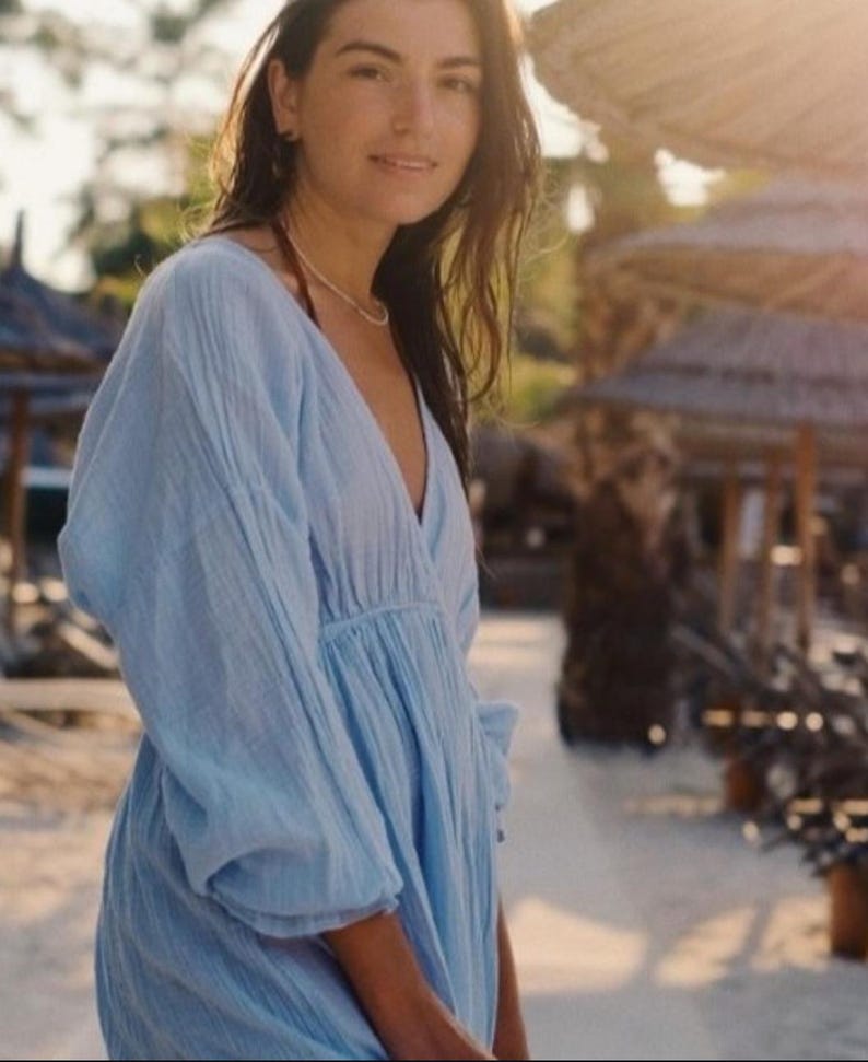 May include: A light blue, long-sleeved dress with a V-neck. The dress has a gathered waist and a flowing skirt. The fabric appears to be lightweight and textured. The dress is worn outdoors in a sunny environment.