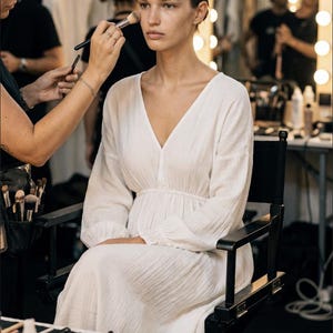 May include: A model in a white dress is having makeup applied in a backstage setting. The dress has a V-neck and long sleeves. Makeup palettes, brushes, and other beauty products are visible on a table in the foreground.