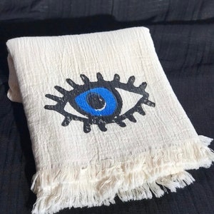 May include: A white cotton throw blanket with a blue evil eye design. The blanket has a fringe border.