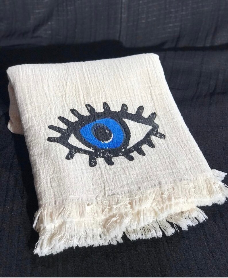 May include: A white cotton throw blanket with a blue evil eye design. The blanket has a fringe border.