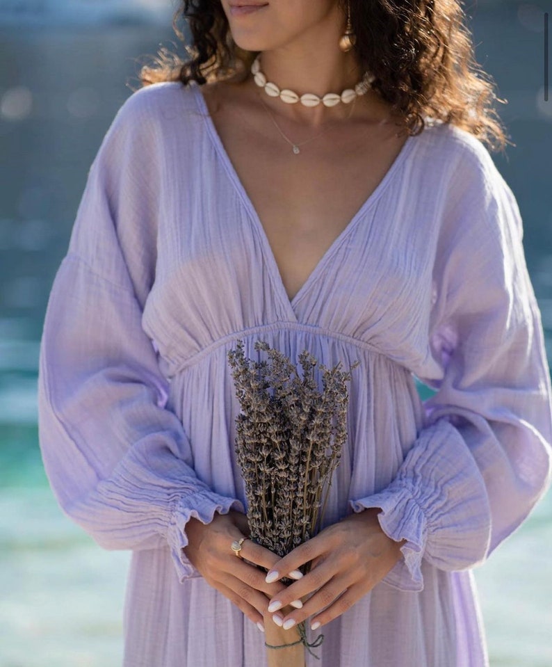 May include: A light purple dress with long sleeves and a V-neck. The dress is gathered at the waist and has ruffled cuffs. A bouquet of dried lavender is held in front of the dress. A shell necklace and gold earrings are also visible.