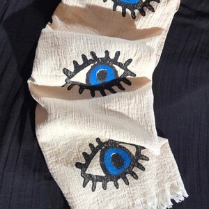May include: A white cotton scarf with a fringe border and three blue evil eye designs. The eyes are outlined in black and have black eyelashes.