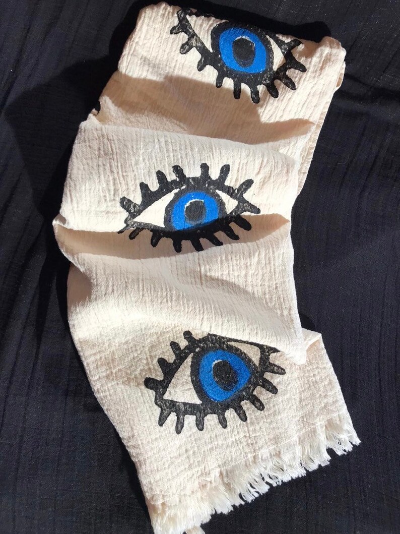 May include: A white cotton scarf with a fringe border and three blue evil eye designs. The eyes are outlined in black and have black eyelashes.