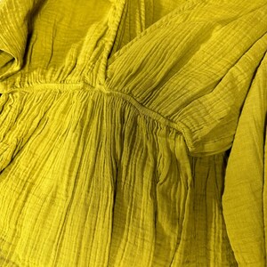 May include: Close-up of a mustard-colored, textured top with a V-neckline. The fabric appears to be a lightweight, crinkled material, possibly cotton or linen. The top has a gathered waist and short sleeves, suggesting a casual style.