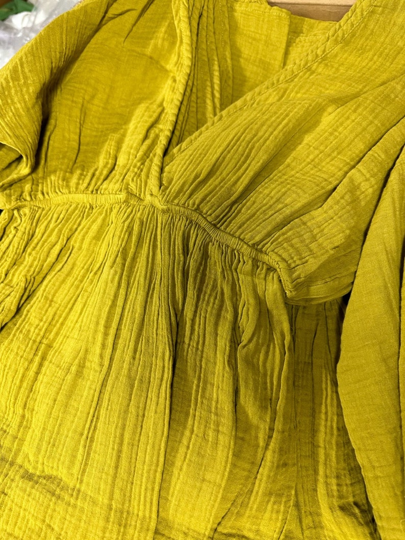 May include: Close-up of a mustard-colored, textured top with a V-neckline. The fabric appears to be a lightweight, crinkled material, possibly cotton or linen. The top has a gathered waist and short sleeves, suggesting a casual style.