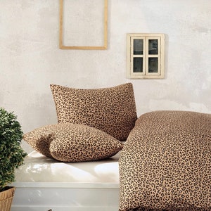May include: A tan and brown leopard print bedding set with two matching pillowcases. The bedding is on a white bed frame with a white wall in the background.