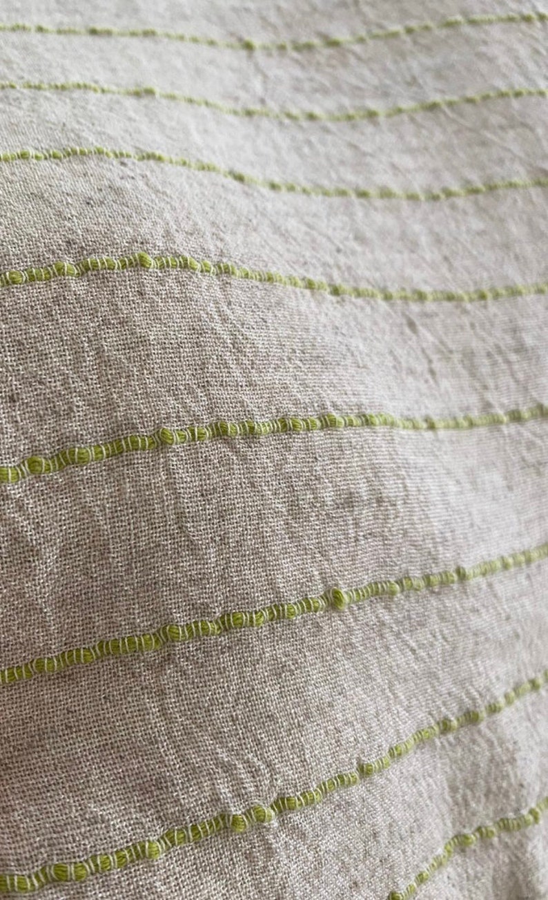 Puede incluir: Primer plano de una tela beige claro con finas rayas verdes. La tela tiene una textura tejida y un brillo sutil.