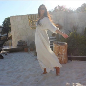 May include: A woman wearing a flowing white dress with long sleeves and a V-neck. The dress is ankle-length and appears to be made of a lightweight fabric. She is standing on a sandy surface, with a stone wall and greenery in the background.