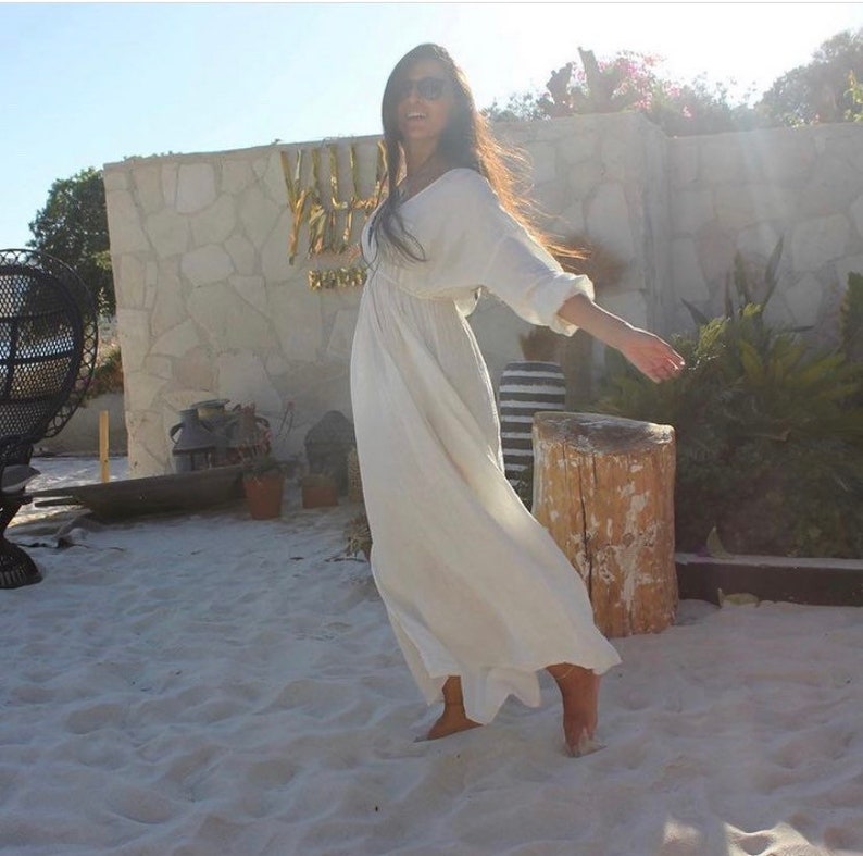 May include: A woman wearing a flowing white dress with long sleeves and a V-neck. The dress is ankle-length and appears to be made of a lightweight fabric. She is standing on a sandy surface, with a stone wall and greenery in the background.