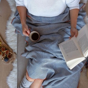 May include: A person is sitting on a couch with a blue and white patterned throw blanket. They are holding a cup of coffee and a book. The blanket has a fringe border.