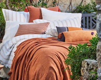 Könnte beinhalten: Ein Bett mit einer weißen und braunen gestreiften Bettdecke und einer rostigen Wolldecke. Das Bett ist mit mehreren Kissen dekoriert, darunter ein braunes gestreiftes Kissen, ein weißes Kissen mit einem braunen Streifen und ein rostiges Kissen mit Quasten.
