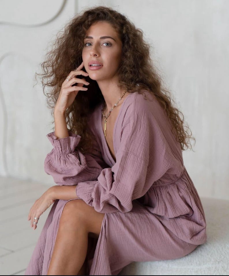 May include: A mauve-colored dress with long sleeves and a deep V-neck. The dress features a cinched waist and a flowing skirt. The sleeves have ruffled cuffs. A gold necklace is visible. The person is sitting on a white surface.
