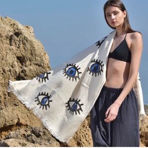 May include: A white beach towel with a fringe border and blue and black eye designs. The towel is draped over a person standing on a beach.