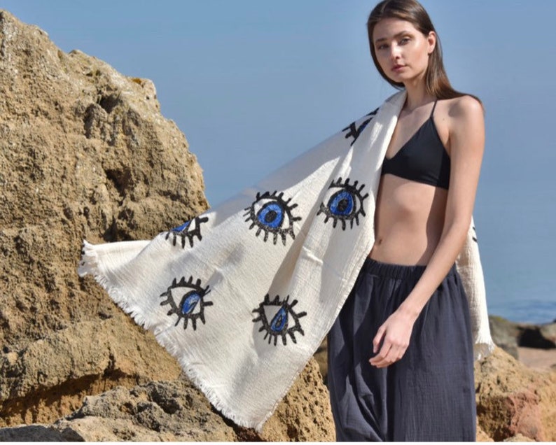May include: A white beach towel with a fringe border and blue and black eye designs. The towel is draped over a person standing on a beach.