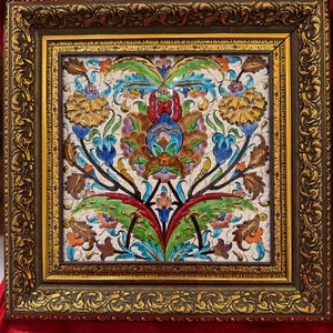 May include: A framed, square ceramic tile with a floral design. The tile features a white background with colorful flowers in shades of blue, green, red, and gold. The frame is ornate, with a dark brown finish and gold accents.