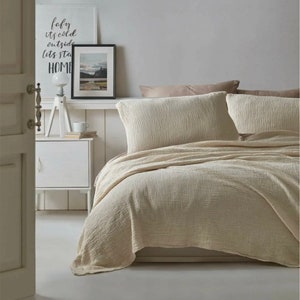 May include: A beige waffle-textured duvet cover and two matching pillowcases on a bed with a white headboard. The bed is in a bedroom with white walls and a white nightstand. A framed print with the text "baby it's cold outside let's stay home" is on the wall behind the bed.
