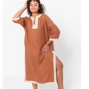 May include: A long, brown linen dress with a white trim around the neckline and sleeves. The dress has a side slit and a relaxed fit.