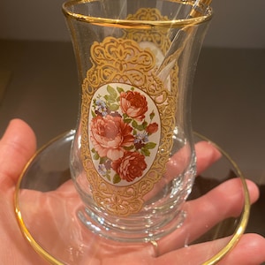May include: A clear glass teacup with a gold rim and a floral design. The cup is sitting on a matching saucer with a gold rim.