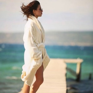 May include: A woman wearing a white robe stands on a wooden dock overlooking a body of water. The robe has a long, flowing design and is tied at the waist.