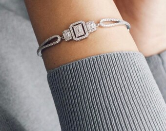 May include: A silver bracelet with a square-shaped diamond centerpiece and smaller diamonds on either side. The bracelet is on a person's wrist.