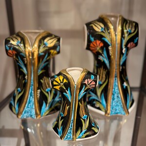 Puede incluir: Tres jarrones decorativos de cerámica con forma de túnica. Los jarrones son negros con ribetes dorados y diseños florales coloridos en azul, naranja y amarillo. Se exhiben en una estante de vidrio.