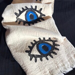 May include: A white cotton towel with a fringe border features three blue evil eye designs. The eyes are outlined in black and have white pupils.