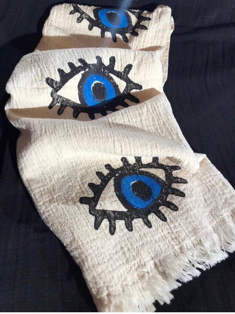 May include: A white cotton towel with a fringe border features three blue evil eye designs. The eyes are outlined in black and have white pupils.
