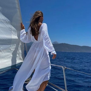 May include: A person stands on a sailboat, wearing a long, flowing white dress with long sleeves and a low back. The dress billows in the wind against a backdrop of a bright blue sky and ocean. A white sail is visible.