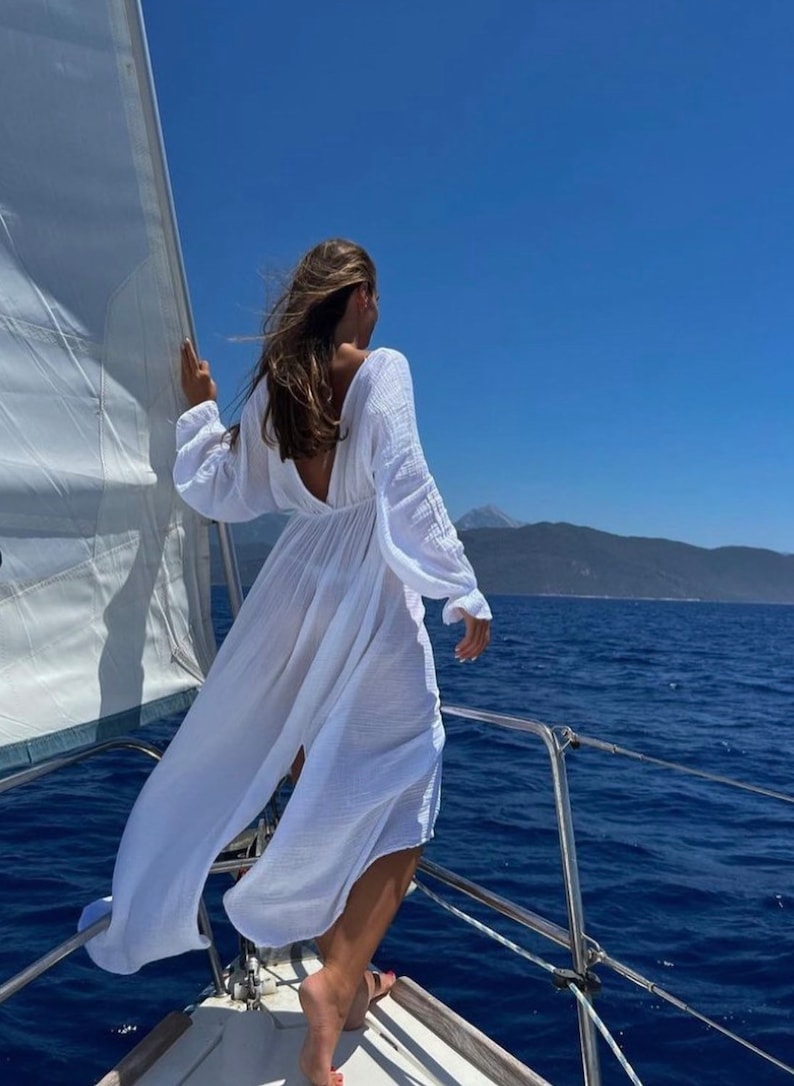May include: A person stands on a sailboat, wearing a long, flowing white dress with long sleeves and a low back. The dress billows in the wind against a backdrop of a bright blue sky and ocean. A white sail is visible.