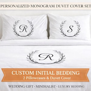 May include: White duvet cover set with monogrammed initials. The set includes a duvet cover and two pillowcases, each featuring a black initial within a laurel wreath design. The text "PERSONALIZED MONOGRAM DUVET COVER SET" is at the top.