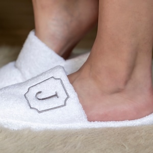 Puede incluir: Pantuflas blancas de felpa con una inicial "J" bordada en gris en un marco decorativo.
