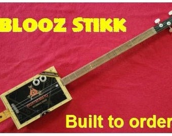 May include: A cigar box guitar with a wooden body and a black finish. The guitar has a single string and is labeled "BLOOZ STIKK". The text "Built to order" is also visible in the image.