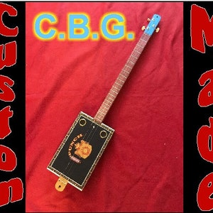 Puede incluir: Una guitarra de caja de puros hecha a medida con un cuerpo negro y un ribete de madera clara. La guitarra tiene una sola cuerda y una pala azul. La guitarra está etiquetada como "C.B.G." y "Custom Made".