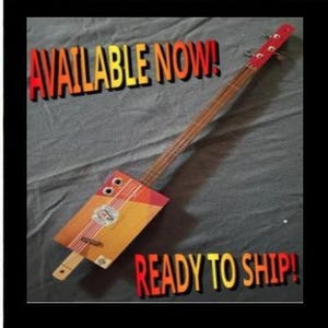 May include: A three-string cigar box guitar with a rectangular wooden body featuring a red, orange, and yellow label with white stripes. The neck is long and thin with a red headstock. The text AVAILABLE NOW! and READY TO SHIP! is visible.