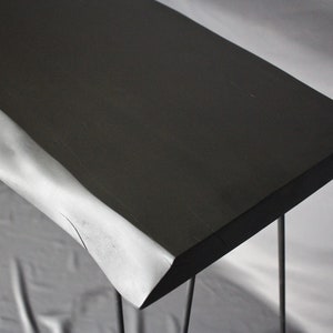 Könnte beinhalten: Ein schwarzer Holz-Konsolentisch mit einer Live-Edge-Oberfläche und dünnen Metallbeinen. Der Tisch steht auf einer weißen Oberfläche.
